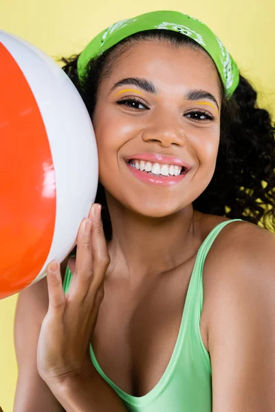 Happy african american young woman in green kerchief holding inflatable ball isolated on yellow — Stock Photo
