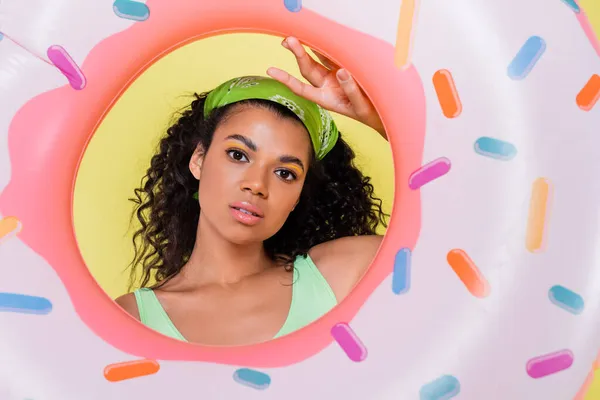 Jeune afro-américaine jeune femme en mouchoir vert tenant anneau gonflable isolé sur jaune — Photo de stock