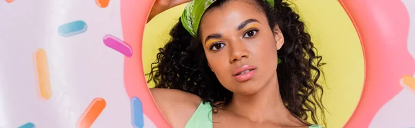 Joven afroamericana joven en pañuelo verde sosteniendo anillo inflable aislado en amarillo, bandera - foto de stock