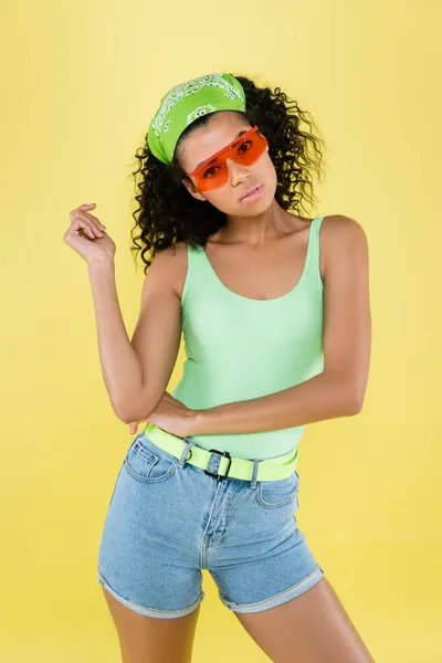 Jovem afro-americana jovem mulher em lenço verde e óculos de sol laranja isolado no amarelo — Fotografia de Stock