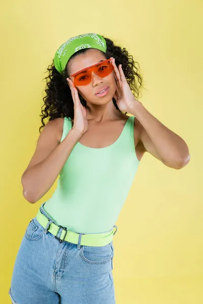 Jeune femme afro-américaine en mouchoir vert ajustant des lunettes de soleil orange isolées sur jaune — Photo de stock