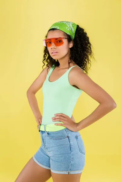 Junge afrikanisch-amerikanische junge Frau in grünem Halstuch und Sonnenbrille, die Hände auf den Hüften isoliert auf gelb — Stockfoto