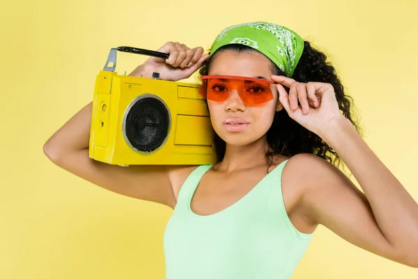 Joven mujer afroamericana en pañuelo y gafas de sol sosteniendo boombox aislado en amarillo - foto de stock