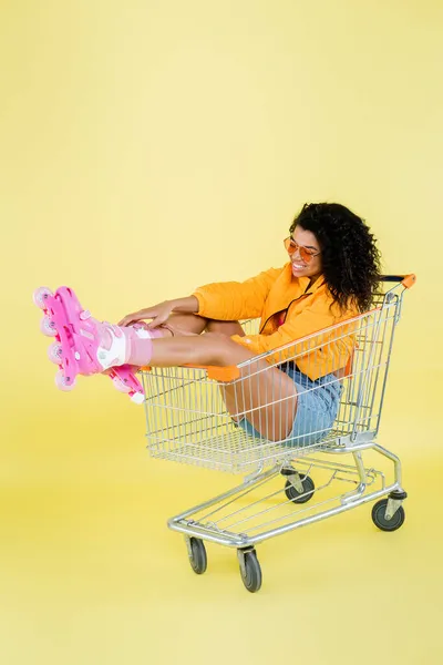 Happy african american young woman in pink roller skates sitting in shopping cart on yellow — Stock Photo