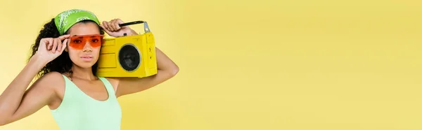 Young african american young woman in kerchief and sunglasses holding boombox isolated on yellow, banner — Stock Photo