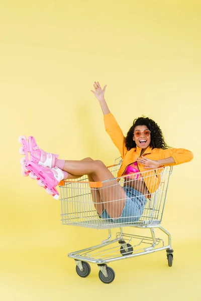 Eccitata afroamericana giovane donna in pattini a rotelle rosa seduta nel carrello della spesa su giallo — Foto stock