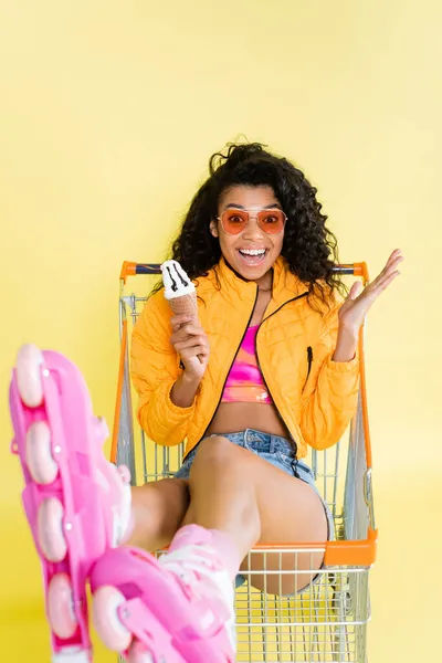 Donna afroamericana stupita in pattini a rotelle rosa con cono gelato mentre seduto nel carrello della spesa su giallo — Foto stock