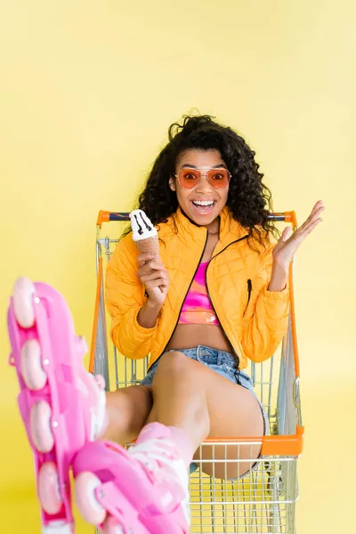 Mujer afroamericana sorprendida en patines rosados con cono de helado mientras está sentada en el carrito de la compra en amarillo - foto de stock