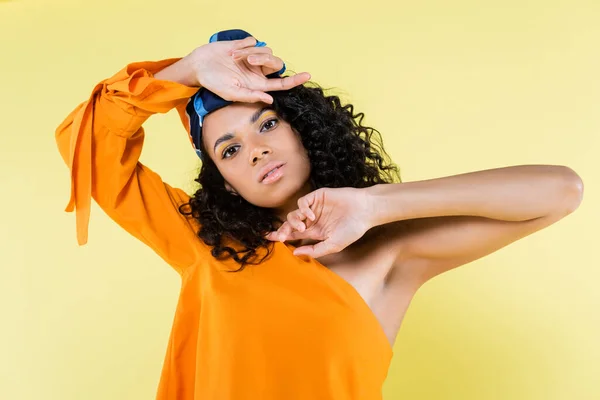 Joven africano americano mujer en la cabeza bufanda posando mientras mira la cámara aislado en amarillo - foto de stock