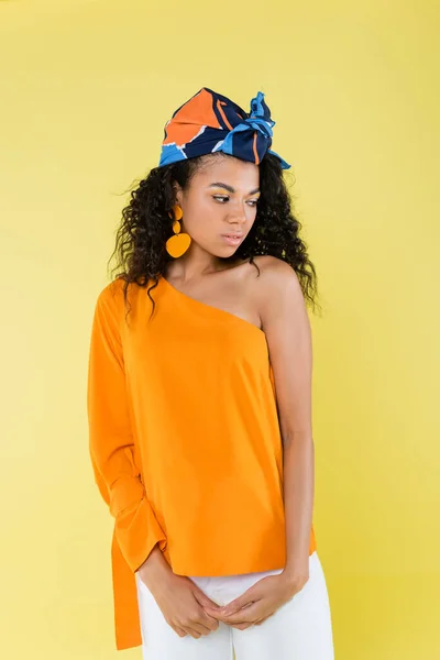 Curly african american woman in kerchief and orange earring looking away isolated on yellow — Stock Photo