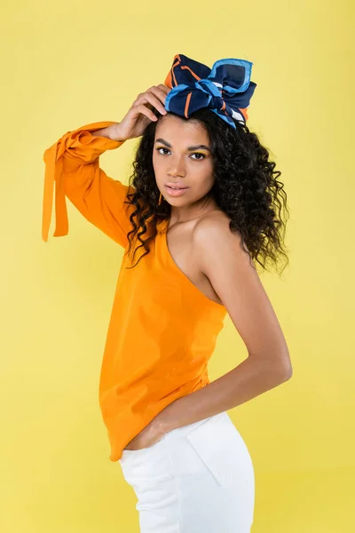 Curly african american woman in headscarf posing with hand on hip isolated on yellow — Stock Photo