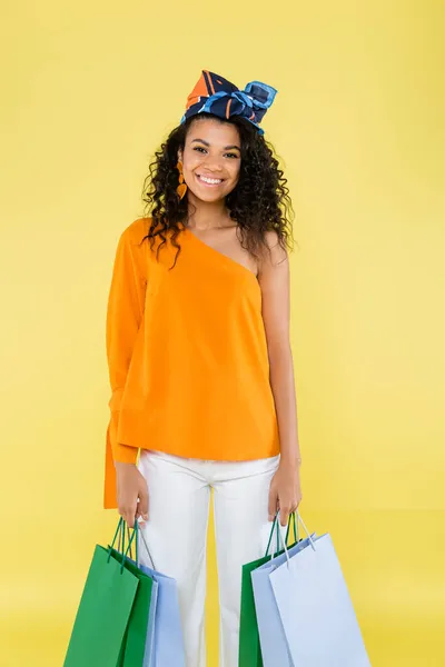 Glückliche afrikanisch-amerikanische Frau mit nackter Schulter, die Einkaufstaschen isoliert auf gelb hält — Stockfoto