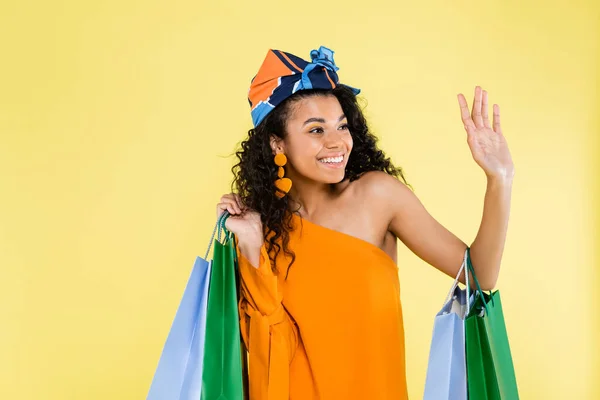 Щаслива афроамериканка з голим плечем тримає сумки для покупок, махаючи рукою ізольовано на жовтому — стокове фото