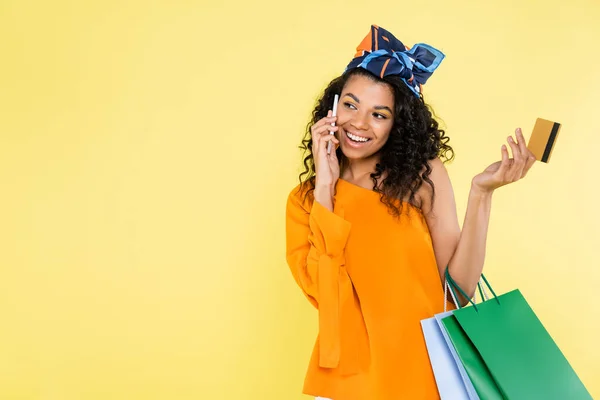 Heureuse femme afro-américaine parlant sur téléphone portable tout en tenant carte de crédit et sacs à provisions isolés sur jaune — Photo de stock