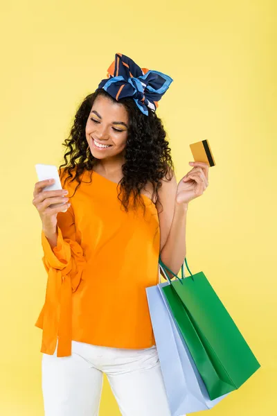 Mulher americana africana alegre segurando celular, cartão de crédito e sacos de compras isolados no amarelo — Fotografia de Stock