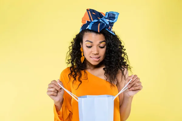 Curious african american woman biting lip while looking at shopping bag isolated on yellow — Stock Photo