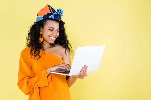 Sorrindo mulher afro-americana usando laptop isolado no amarelo — Fotografia de Stock