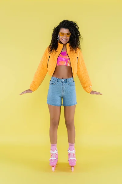 Pleine longueur de positive et frisée afro-américaine jeune femme en patins à roulettes roses sur jaune — Photo de stock