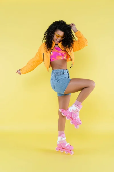 Comprimento total de feliz e encaracolado Africano americano jovem mulher em patins de rolo rosa no amarelo — Fotografia de Stock