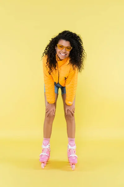 Comprimento total de alegre e encaracolado Africano americano jovem mulher em patins de rolo rosa no amarelo — Fotografia de Stock