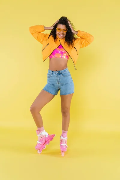 Pleine longueur de jeune femme africaine américaine heureuse et bouclée en patins à roulettes ajustant les cheveux sur jaune — Photo de stock