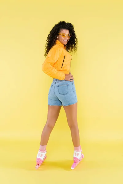 Pleine longueur de positive et frisée afro-américaine jeune femme en veste gonflante et patins à roulettes roses sur jaune — Photo de stock