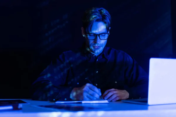 Young programmer writing on notebook near laptop on table on black background — Stock Photo