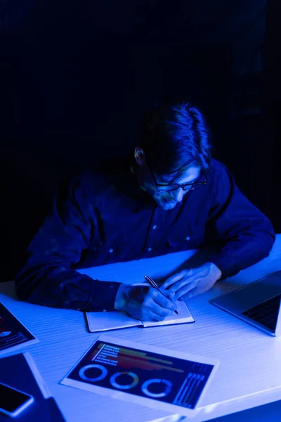 Programador en anteojos escribiendo en cuaderno cerca de papeles y dispositivos en mesa aislados en negro - foto de stock