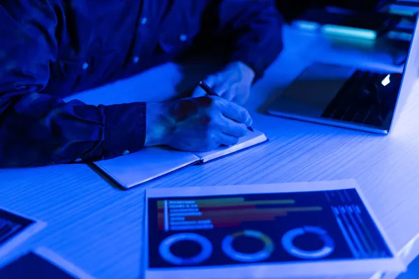 Cropped view of programmer writing on notebook near laptop on table on black background — Stock Photo