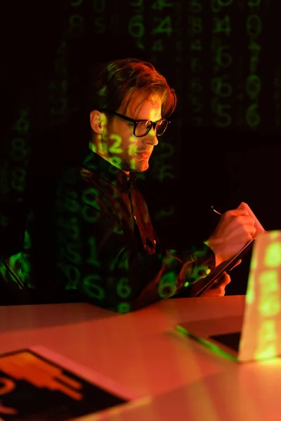 Programmer in eyeglasses writing on notebook near blurred laptop and reflection of computer code on black background — Stock Photo