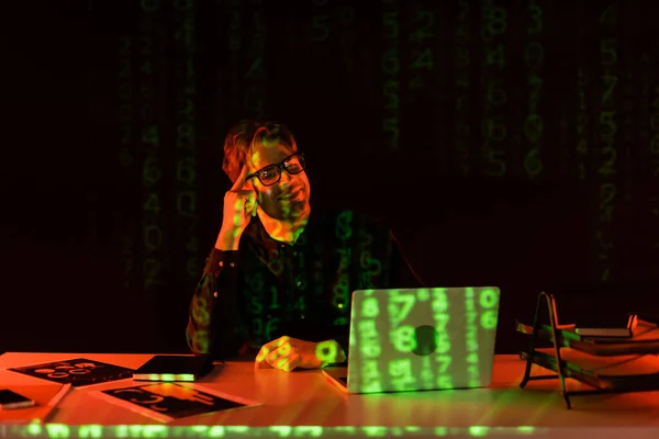 Smiling programmer in eyeglasses looking at laptop near reflection of computer code on black background — Stock Photo