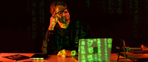 Programador sonriente en anteojos mirando el ordenador portátil con el reflejo del código informático cerca de papeles sobre fondo negro, bandera - foto de stock