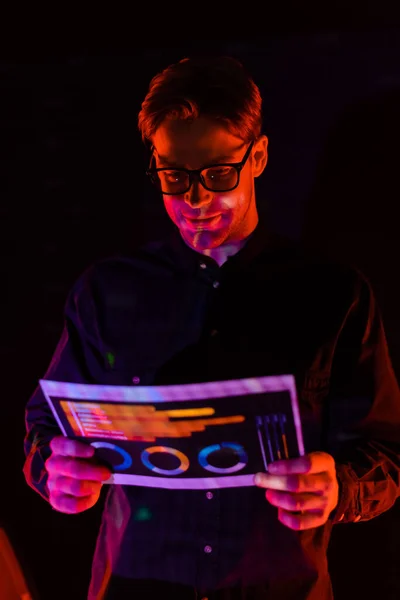 Young programmer in eyeglasses holding paper with charts isolated on black — Stock Photo