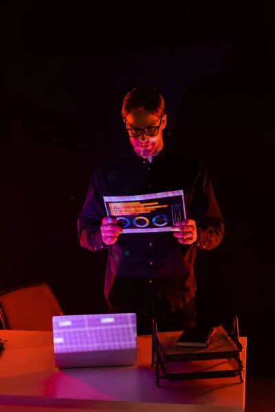 Young programmer holding paper with charts near laptop on table isolated on black — Stock Photo