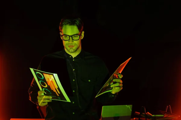 Programmer holding papers with graphs near laptop on black background with lighting — Stock Photo