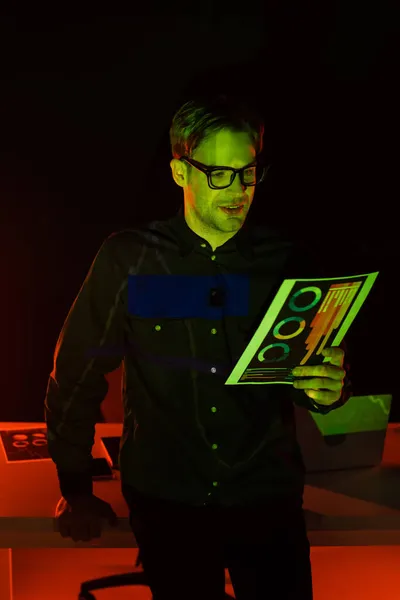 Усміхнений програміст тримає документ з графіками біля ноутбука з відображенням на чорному тлі — стокове фото