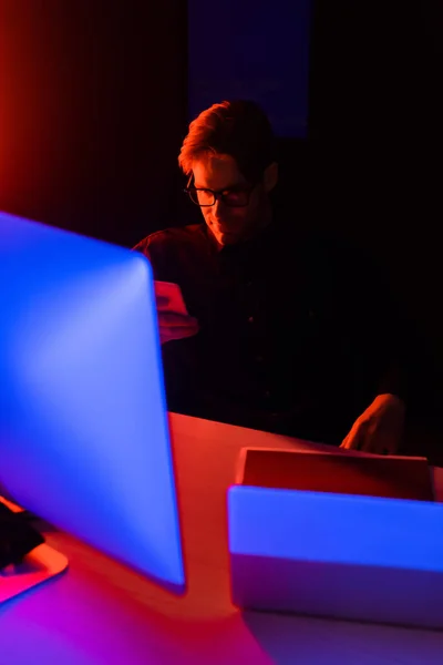 Programador en anteojos usando smartphone cerca de computadoras en mesa sobre fondo negro - foto de stock