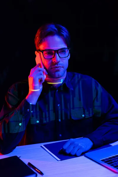 Programmer in eyeglasses talking on cellphone near laptop and paper folder isolated on black — Stock Photo