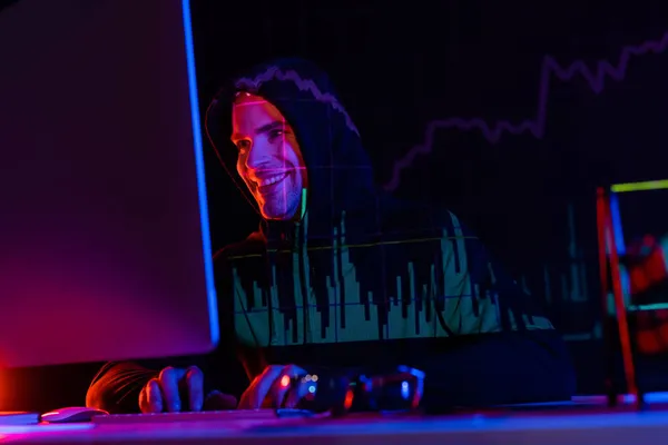Hackeador sonriente usando computadora cerca de anteojos y reflejo de gráficos sobre fondo negro - foto de stock