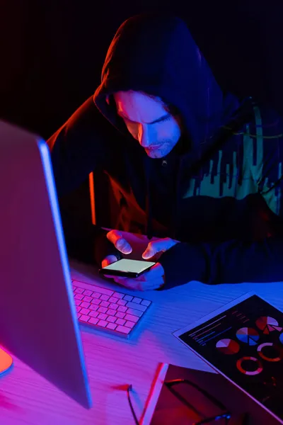 Hacker mit Reflexion von Diagrammen mit Smartphone in der Nähe von Computer und Papiere isoliert auf schwarz — Stockfoto