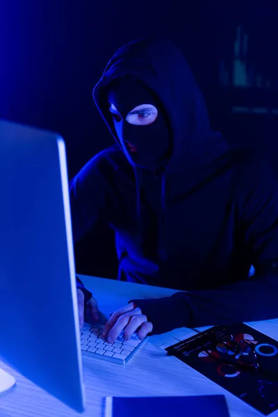 Hacker en pasamontañas usando computadora cerca de papeles en la mesa sobre fondo negro - foto de stock