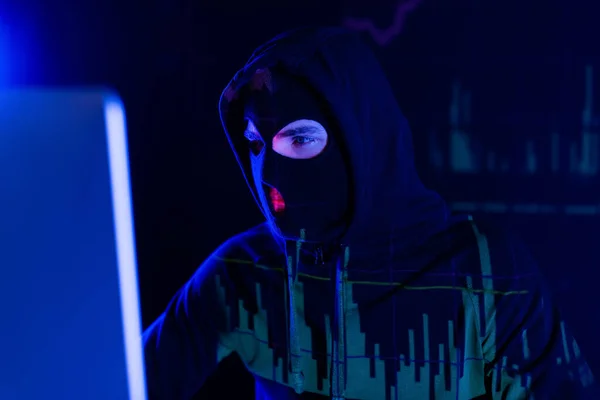 Hacker in balaclava with charts reflection looking at blurred computer monitor on black background — Stock Photo