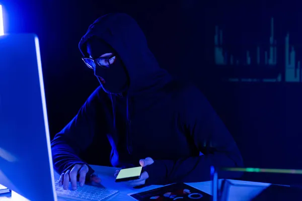 Hacker in passamontagna utilizzando computer e smartphone su sfondo nero con grafici di riflessione — Foto stock