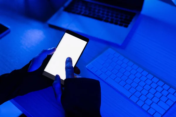 Vista recortada del programador utilizando el teléfono inteligente cerca del ordenador portátil y el teclado de la computadora en la mesa - foto de stock