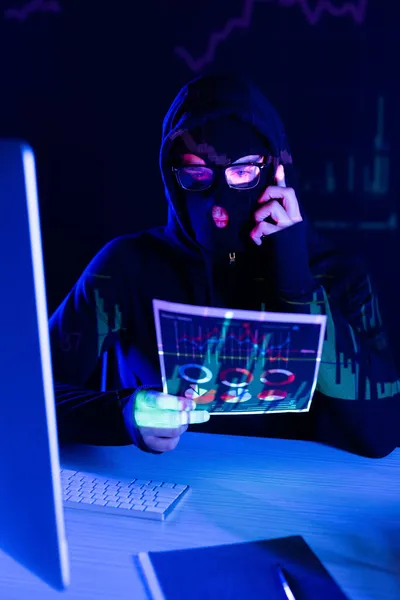 Hacker in balaclava talking on smartphone and holding paper near computer on black background — Stock Photo