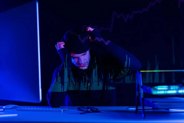 Young hacker wearing balaclava near computer and papers on black background with charts reflection — Stock Photo