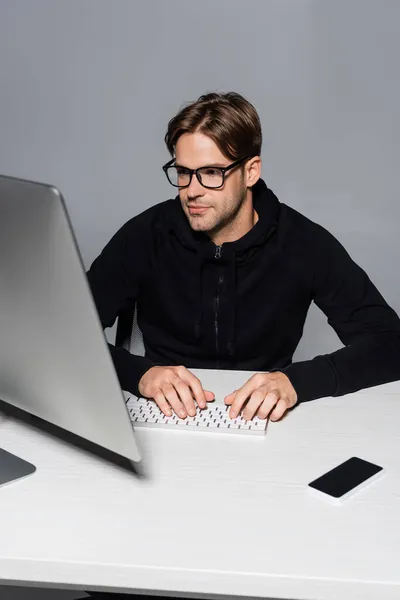 Programador en anteojos con ordenador cerca de smartphone aislado en gris - foto de stock