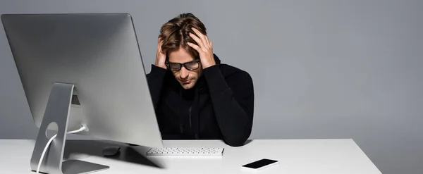 Displeased programmer in eyeglasses sitting near computer and smartphone isolated on grey, banner — Stock Photo
