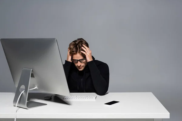 Programador molesto sentado cerca de la computadora y el teléfono celular en la mesa aislado en gris - foto de stock