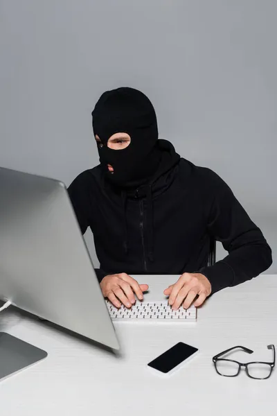 Hacker en pasamontañas usando computadora cerca de smartphone y anteojos aislados en gris - foto de stock